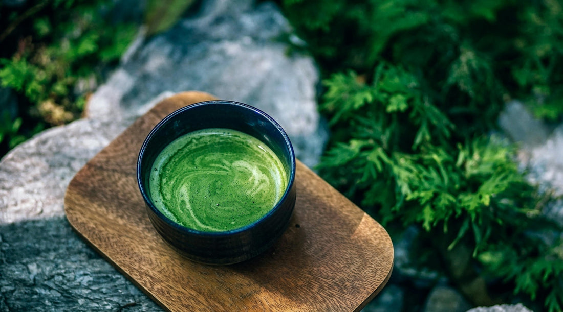 A bowl of freshly whisked matcha tea with a frothy swirl, placed on a wooden tray on a rocky surface with greenery in the background