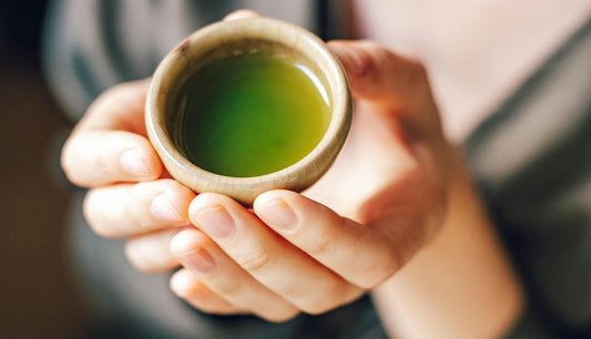 Close-up of hands gently holding a small ceramic cup filled with vibrant green matcha tea, symbolizing warmth, relaxation, and mindfulness.