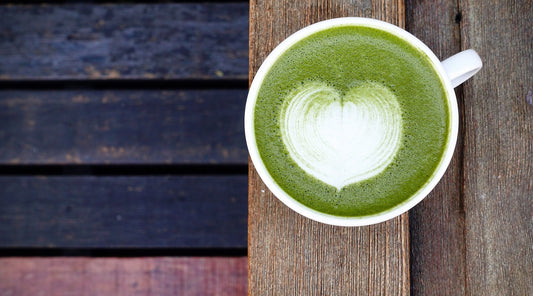 Top view of a matcha latte in a white cup with heart-shaped latte art, placed on a rustic wooden table. The vibrant green color of the matcha contrasts with the earthy background, creating a cozy and inviting scene.