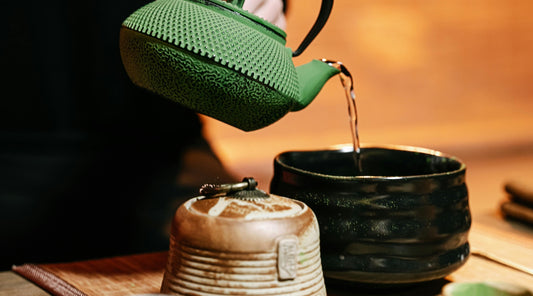 Close-up of a green teapot pouring hot water into a traditional Japanese tea bowl, set against a warm background. The scene highlights the ritual of preparing tea with authentic Japanese tea ware.